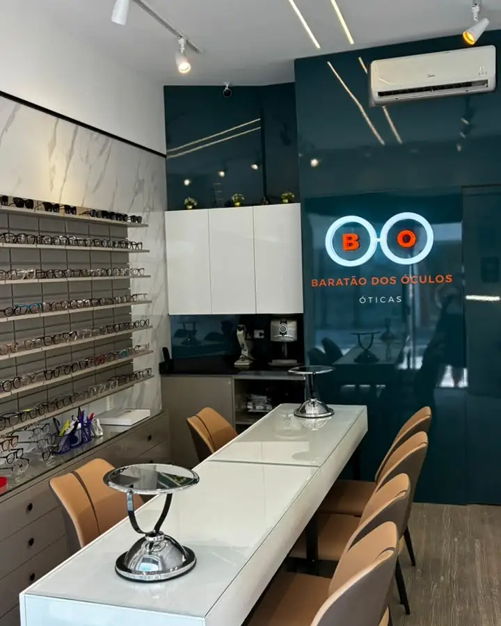Interior da Óticas Baratão dos Óculos: mesa de atendimento branca, cadeiras e parede verde com a logo BO iluminada